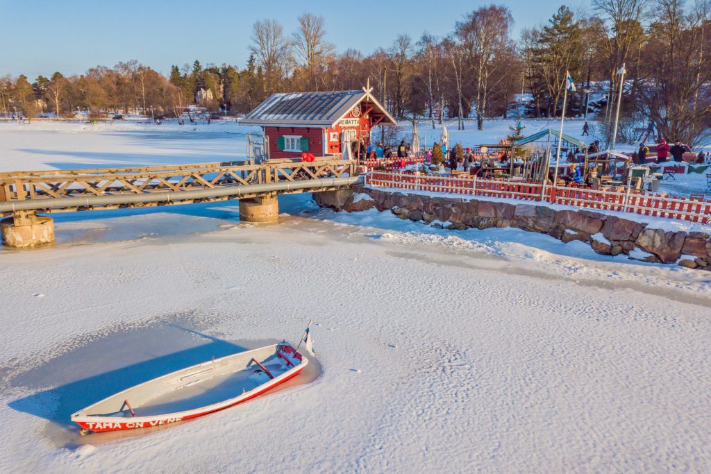 A view from the the frozen water looking towards land and Cafe Regatta in winter. Everything is covered by a light dusting of snow, a boat is sitting in the ice in the bottom left of the photo, and the trees of Sibelius Park cover the horizon in the background. People are queuing to get into the cafe.