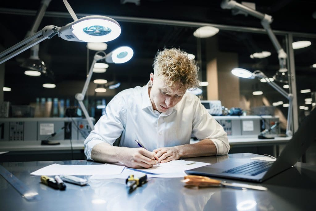 Sat facing the camera, a man at a workstation writes notes on paper, his laptop in front and to the left of him, scissors, pliers and other small tools scattered on the table and an angle-poise lamp to his right illuminating the workspace
