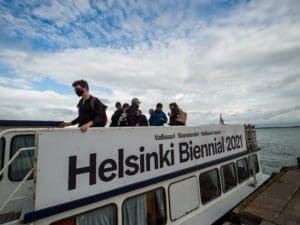 90 Day Finn participants are waiting to take their seats on top of a ferry to Helsinki Biennial 2021, held Vallisaari island. The boat is at dock with a partially cloudy and blue sky behind them.