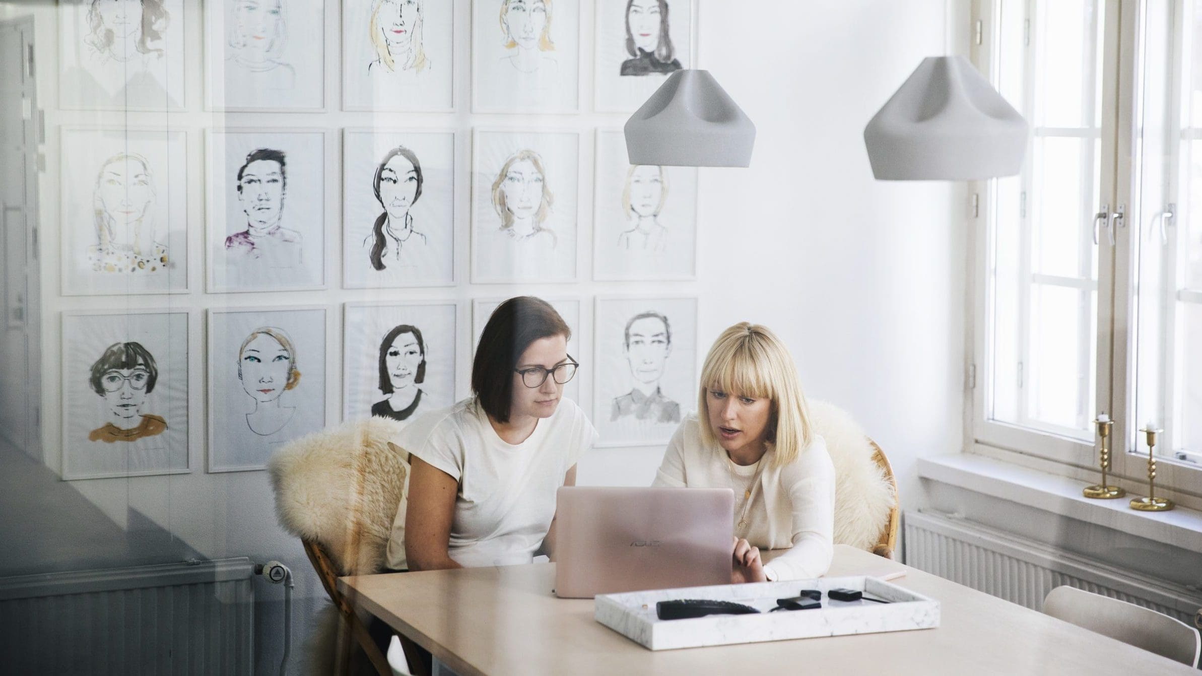 Viewed from behind an office window which shows slight reflections, two women are sitting at a table discussing something that is on a laptop in front of them. The office wall behind them is covered in stylised, illustrated portraits.