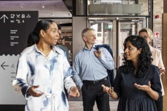 A group of international people walking and talking, there are two women in front and a man and women in the back. They all have different ages and backgrounds. They walk inside an event venue.