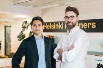 The two partners from NordicNinja (men) are standing and looking at the camera. Behind them there is the Helsinki Partners logo and the Helsinki Partners office background. One of the man is from Japan and is wearing a black suit and a blue shirt, the other man is from Finland and he wears a white shirt.