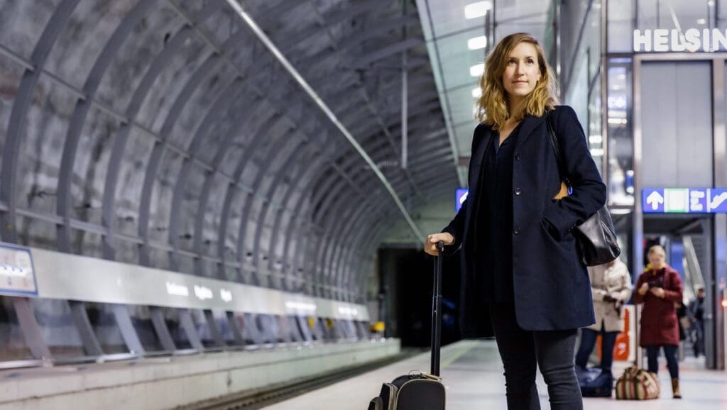 Standing in the huge underground tunnel that is the Helsinki airport tain station, a woman wearing a long blue coat stands at the platform waiting for her train into Helsinki. On the left are the train tracks and on the right are the lifts and escalators to the airport.