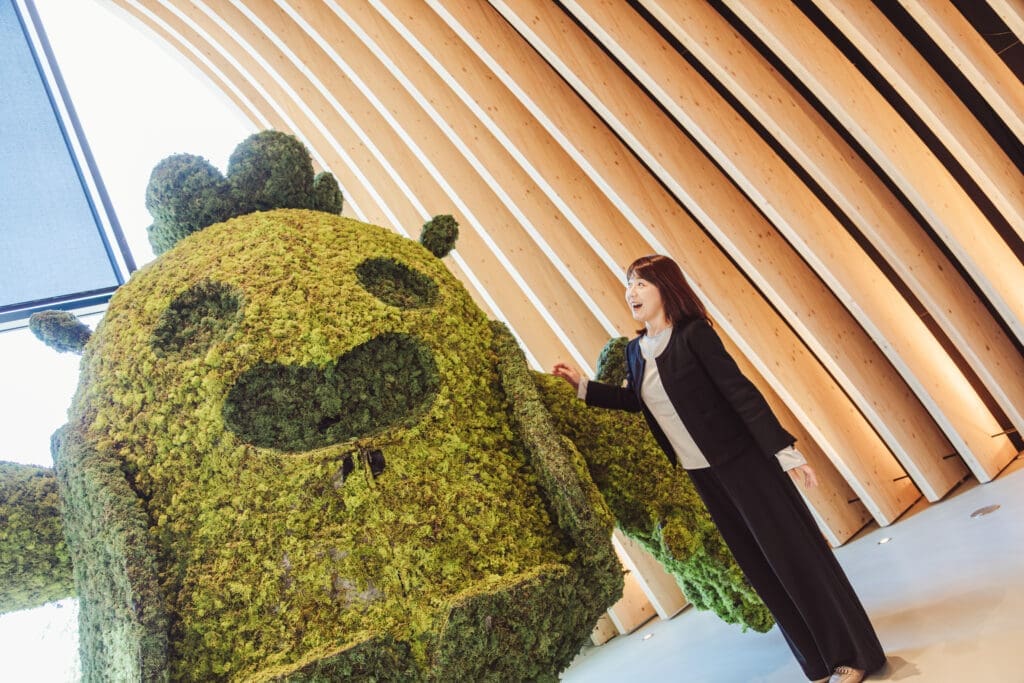 Woman stands next to a large game character made of out of moss mouth open in excitement 